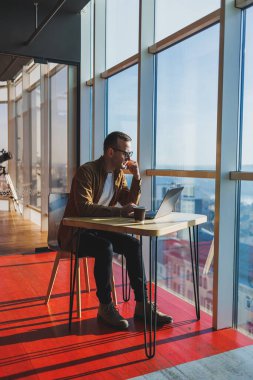 A balanced man in glasses and casual clothes works on a laptop from a cozy workspace. A successful freelancer works remotely.