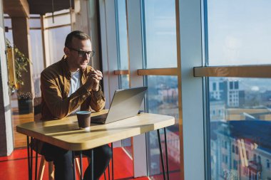 A balanced man in glasses and casual clothes works on a laptop from a cozy workspace. A successful freelancer works remotely.