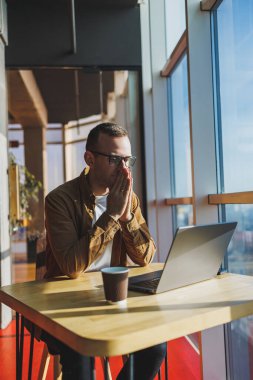 A balanced man in glasses and casual clothes works on a laptop from a cozy workspace. A successful freelancer works remotely.