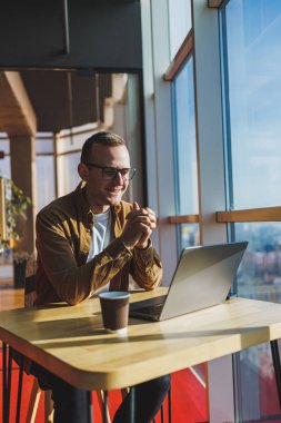 A balanced man in glasses and casual clothes works on a laptop from a cozy workspace. A successful freelancer works remotely.