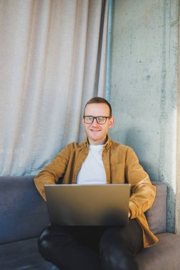 Young male worker in casual clothes and glasses looking at laptop while sitting on sofa with laptop in bright modern workspace. A successful freelancer works remotely online in a workspace
