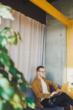 Young male worker in casual clothes and glasses looking at laptop while sitting on sofa with laptop in bright modern workspace. A successful freelancer works remotely online in a workspace