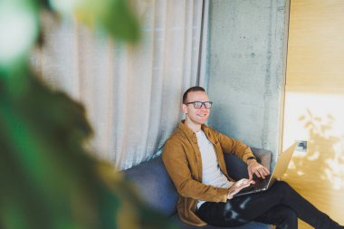 Young male worker in casual clothes and glasses looking at laptop while sitting on sofa with laptop in bright modern workspace. A successful freelancer works remotely online in a workspace