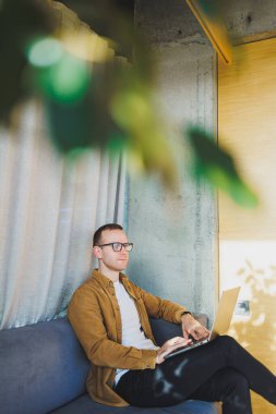 Young male worker in casual clothes and glasses looking at laptop while sitting on sofa with laptop in bright modern workspace. A successful freelancer works remotely online in a workspace