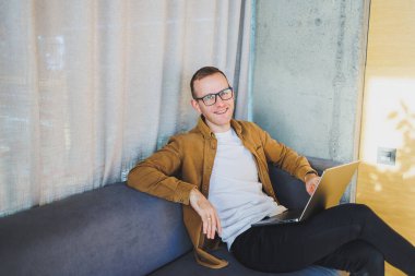 Young male worker in casual clothes and glasses looking at laptop while sitting on sofa with laptop in bright modern workspace. A successful freelancer works remotely online in a workspace