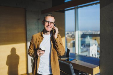 Smiling male worker in casual clothes looking away while talking on mobile phone and drinking coffee during break in modern creative workspace