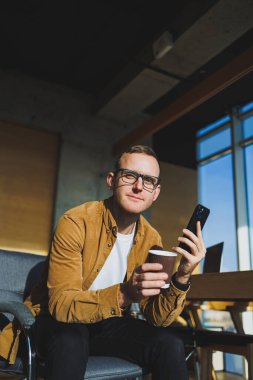 Smiling male worker in casual clothes looking away while talking on mobile phone and drinking coffee during break in modern creative workspace