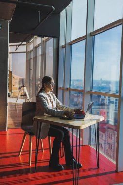 Young content worker in casual clothes sitting at a desk and using a netbook while working on the background of a spacious office. Business woman in the office works sitting near the window