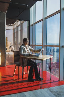 Young content worker in casual clothes sitting at a desk and using a netbook while working on the background of a spacious office. Business woman in the office works sitting near the window