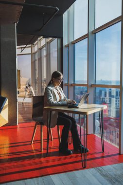 Young content worker in casual clothes sitting at a desk and using a netbook while working on the background of a spacious office. Business woman in the office works sitting near the window