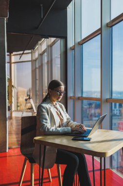 Young content worker in casual clothes sitting at a desk and using a netbook while working on the background of a spacious office. Business woman in the office works sitting near the window