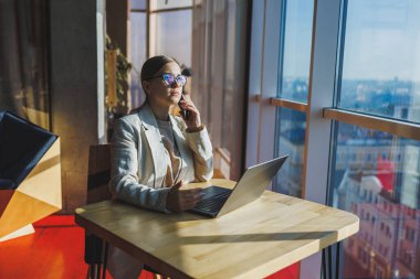 Cheerful young woman in casual wear having telephone conversation sitting at working place near laptop in office, smiling female freelancer calling to colleague for share good news about business