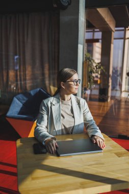 Young content worker in casual clothes sitting at a desk and using a netbook while working on the background of a spacious office. Business woman in the office works sitting near the window