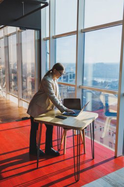 Cheerful young female freelancer in casual clothes standing at a wooden table with a netbook and writing notes while working on a project. Modern young business lady in the office