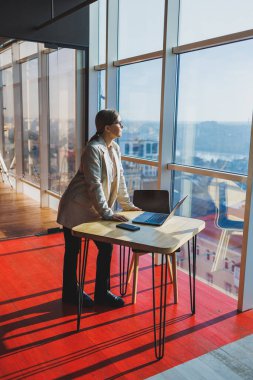 Cheerful young female freelancer in casual clothes standing at a wooden table with a netbook and writing notes while working on a project. Modern young business lady in the office
