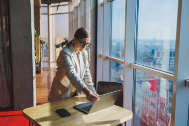 Cheerful young female freelancer in casual clothes standing at a wooden table with a netbook and writing notes while working on a project. Modern young business lady in the office