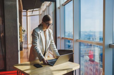 Cheerful young female freelancer in casual clothes standing at a wooden table with a netbook and writing notes while working on a project. Modern young business lady in the office