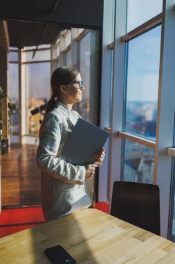 Cheerful young female freelancer in casual clothes standing at a wooden table with a netbook and writing notes while working on a project. Modern young business lady in the office