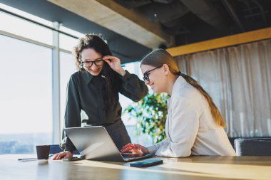 Smiling female colleagues working on new project at laptop, working in modern spacious office with large windows. Woman in glasses and casual clothes in the office.