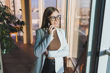Top view of positive woman in casual clothes standing near big window and talking on phone, giving information during remote project. Modern workspace for remote work.