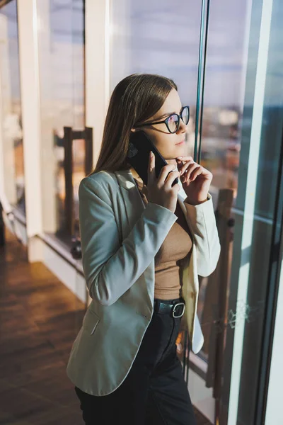 Top view of positive woman in casual clothes standing near big window and talking on phone, giving information during remote project. Modern workspace for remote work.