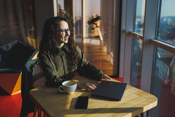 A cute woman works on a laptop in a cafe. Young concentrated brunette woman in glasses sitting at the table near the window drinking coffee. Freelance and remote work. Modern women's lifestyle