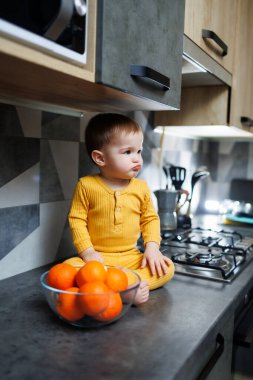 A little boy 1 year old in yellow clothes is sitting in the kitchen on the table with a plate of orange tangerines. Portrait of a cute one-year-old boy and sweet citrus fruits.
