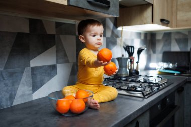 A little boy 1 year old in yellow clothes is sitting in the kitchen on the table with a plate of orange tangerines. Portrait of a cute one-year-old boy and sweet citrus fruits.