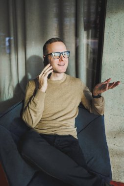 Gözlüklü gülümseyen genç bir adam ofiste dinlenirken telefonla gülümsüyor ve konuşuyor. Genç bir serbest çalışan uzaktan çalışıyor..