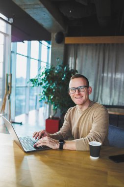 Genç serbest çalışan gözlüklü adam, modern bir çalışma alanında uzaktan kumandalı bir proje üzerinde çalışırken tahta bir masada dizüstü bilgisayar ve kahveyle oturuyor..