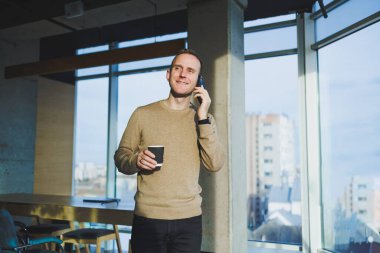 Ofiste dinlenirken gülümseyen, telefonla konuşan ve kahve içen olumlu genç adam. İş sırasında öğle yemeği molası.