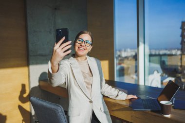 Cute positive female employee with long blond hair in casual clothes looking at phone while working on new business project at table with laptop and gadgets in office