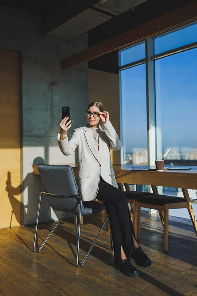 Cute positive female employee with long blond hair in casual clothes looking at phone while working on new business project at table with laptop and gadgets in office