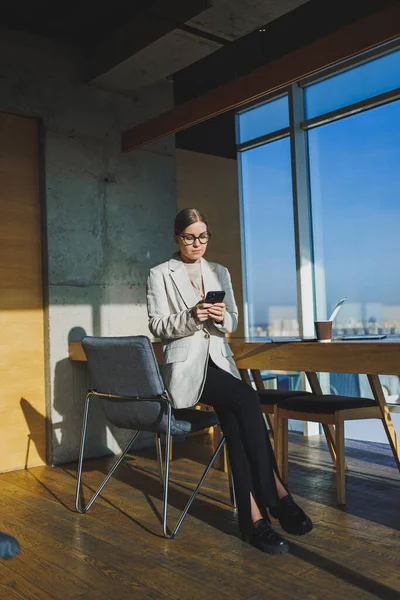 Cute positive female employee with long blond hair in casual clothes looking at phone while working on new business project at table with laptop and gadgets in office