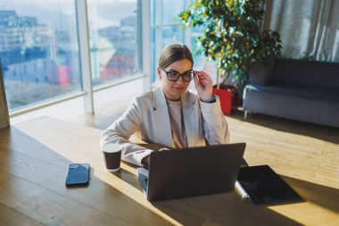 A positive business woman in a jacket is sitting at a table, working on a laptop in a bright office. A young European woman is sitting at a table. Freelance and remote work. Modern women's lifestyle