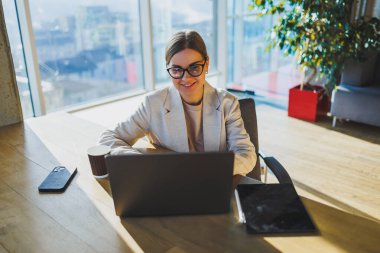 A positive business woman in a jacket is sitting at a table, working on a laptop in a bright office. A young European woman is sitting at a table. Freelance and remote work. Modern women's lifestyle