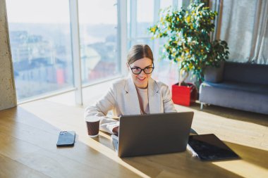 A positive business woman in a jacket is sitting at a table, working on a laptop in a bright office. A young European woman is sitting at a table. Freelance and remote work. Modern women's lifestyle