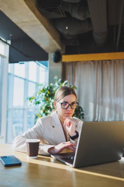 A positive business woman in a jacket is sitting at a table, working on a laptop in a bright office. A young European woman is sitting at a table. Freelance and remote work. Modern women's lifestyle
