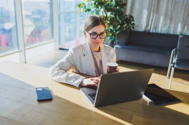 A positive business woman in a jacket is sitting at a table, working on a laptop in a bright office. A young European woman is sitting at a table. Freelance and remote work. Modern women's lifestyle