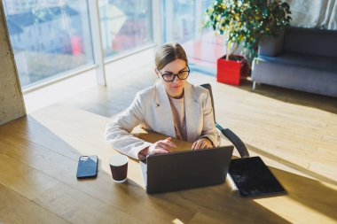A positive business woman in a jacket is sitting at a table, working on a laptop in a bright office. A young European woman is sitting at a table. Freelance and remote work. Modern women's lifestyle
