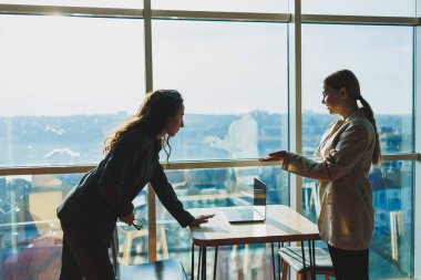 Two business women are standing against the background of a large window in a modern office with a panoramic view of the city. Modern business women work remotely
