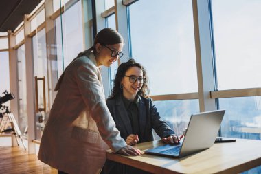 Two business women are looking for something in a laptop. Concept of business cooperation and teamwork. Young smiling women at the desk in the office. Modern successful people