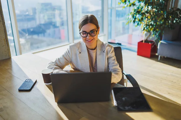A positive business woman in a jacket is sitting at a table, working on a laptop in a bright office. A young European woman is sitting at a table. Freelance and remote work. Modern women's lifestyle