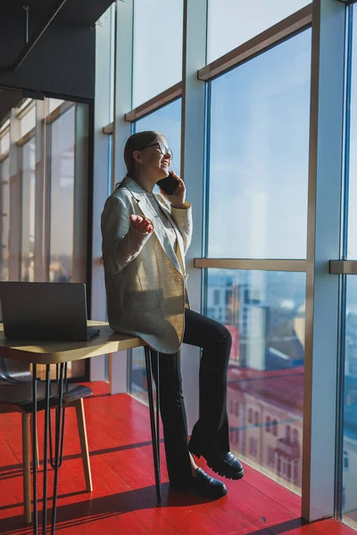 Smiling businesswoman in elegant suit standing in front of window against skyscrapers background, modern spacious office space. Modern office women at work