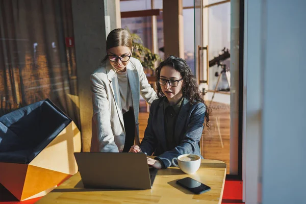 Two business women are looking for something in a laptop. Concept of business cooperation and teamwork. Young smiling women at the desk in the office. Modern successful people