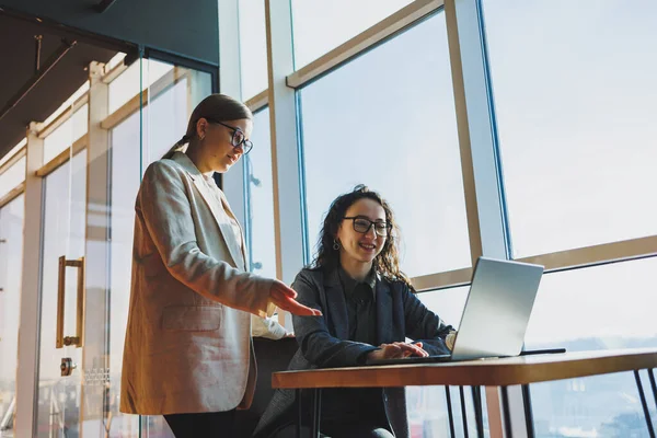 Two business women are looking for something in a laptop. Concept of business cooperation and teamwork. Young smiling women at the desk in the office. Modern successful people