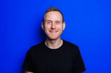 A young man in a black T-shirt on a blue background shows different emotions on his face. Cheerful man with a smile. Place for text. Male beauty