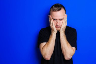 A young man in a black T-shirt on a blue background shows different emotions on his face. Cheerful man with a smile. Place for text. Male beauty