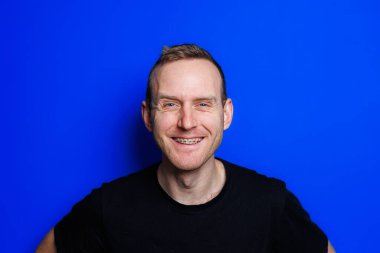 A young man in a black T-shirt on a blue background shows different emotions on his face. Cheerful man with a smile. Place for text. Male beauty