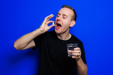 A young man in a black T-shirt on a blue background stands with a thermometer in his hands. An increase in body temperature, feeling sick. Weakened immunity. Selective focus